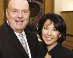 A man with a bald head wearing a suit and tie stands next to a woman with shoulder-length dark hair in a black suit jacket. They are both smiling, standing in an elegant indoor setting adorned with artwork and holding glasses of champagne.