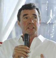 A person with short dark hair is speaking into a microphone. They are wearing a white shirt and standing in front of a backdrop with light and shadows. Their hand is raised, gesturing as they speak passionately about Marin Organic initiatives.