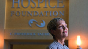 A woman with short gray hair stands in front of a sign that reads "Hospice Foundation" in a well-lit space. She is wearing a gray shirt and looking slightly to the side while smiling softly, navigating the ultimate transition. Two lamps on either side of the sign add a warm glow to the scene.