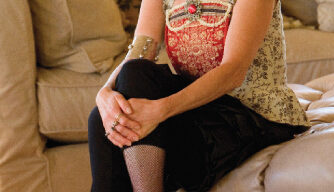 A person with short dark hair sits on a beige sofa, smiling. Dressed in a patterned top, black pants, fishnet stockings, and red high heels, they wear a large bead necklace and a metallic forearm cuff. With hands crossed over their knee, they exude the chic style of Tzeira Sofer.