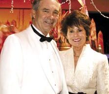 A man and a woman dressed in formal white attire smile for the camera amid a festive background with string lights and colorful decorations at Marin's 12th Annual Valentine's Ball. The man wears a white tuxedo with a black bow tie, while the woman dons a white dress with a black belt.