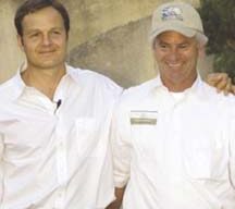 Two men stand side by side, smiling, with one man's arm around the other's shoulder. Both are wearing white shirts, and the man on the right has a name tag and is wearing a beige baseball cap. They are outdoors at the Taste of Marin event, with a beige wall and greenery in the background.