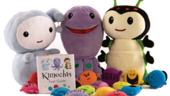 Three stuffed animals from the Kimochis collection are lined up in a row next to a book titled "Kimochis Feel Guide." In front of them, like currents of emotions, colorful plush toys bearing various feelings are scattered playfully.