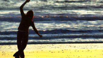 A person is walking along a beach at sunset, silhouetted against the reflective water. They have one arm raised and appear to be enjoying the peaceful scene like a graceful sand dancer. Waves gently roll in the background under the warm, golden light.