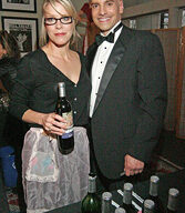 A man in a tuxedo stands next to a woman wearing glasses and a black top, holding a bottle of Mad Housewife Wine. They are in front of a table with several bottles of wine on it, and pictures from an Oscar Party are hanging on the wall in the background.
