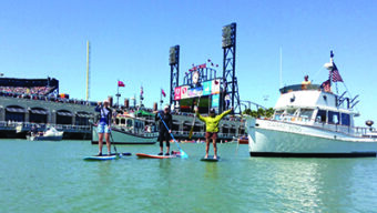 Three people are paddleboarding on water near a large boat and a stadium with a scoreboard and flags. The sunny sky adds to the vibrant atmosphere as onlookers crowd the stadium and the docked boat to watch these popular summer events unfold, offering perfect July activities for all ages.