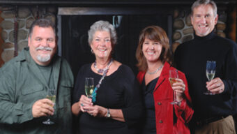 Four smiling people holding glasses of champagne stand side by side in front of a stone fireplace. They appear to be at a celebratory event, dressed in semi-formal attire, including shirts, a black sweater, and a red jacket—perfect holiday hosting inspiration.