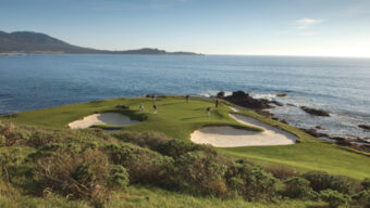 Scenic view of a golf course situated along a coastal cliff. Four golfers are playing on a green surrounded by sand bunkers, with lush greenery in the foreground. The ocean and distant hills are visible under a clear sky, creating an idyllic kingdom for golf enthusiasts.