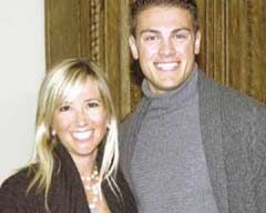 A man and a woman stand closely together, both smiling at the camera. The woman has long blonde hair and is wearing a dark sweater, while the man has short hair and is wearing a grey TSE turtleneck sweater. They are standing in front of a wooden background in Tiburon.