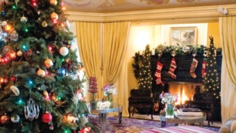 A festive living room is decorated for the holidays with a large, illuminated tree adorned with ornaments. Stockings hang over a fireplace, and wrapped gifts sit underneath the tree. A colorful ceiling mural and cozy seating area add to the warm, celebratory atmosphere.