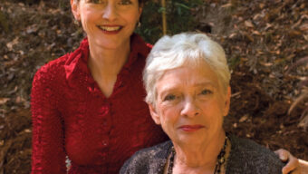 Two women, possibly pen pals, are pictured outdoors. One, standing, has short dark hair and is wearing a red blouse. The other, seated, has short white hair and is dressed in a grey cardigan. They are positioned in front of a natural hillside background with leaves and greenery.