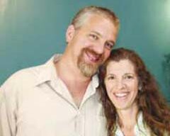 A smiling man and woman standing closely together in front of a teal background create a delightful fusion of warmth and color. The man, with short light hair, a beard, and a light-colored shirt, stands beside the woman with long, wavy hair who is also dressed in a light-colored shirt.