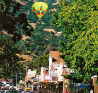 A hot air balloon floats above the small town of Calistoga, with tree-lined streets and mountains in the background. Cars are parked along the sidewalk, and people are walking on the street. The sky is clear and blue, suggesting a sunny day.