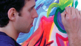 A man with dark curly hair is painting on a canvas filled with vibrant colors including purple, red, yellow, and green. He is holding a paintbrush and appears focused on adding details to the artwork, using art as therapy to express his emotions.