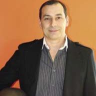 A man with short dark hair and a slight smile is standing against an orange background. He is wearing a dark blazer over a striped shirt, and his right hand is resting on what appears to be a helmet with the logo of Blowtorch Inc.