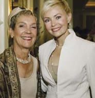 Two women smiling at the camera. The woman on the left has short hair, is wearing a headband, a pearl necklace, and a patterned shawl. The woman on the right has short blonde hair and is wearing a white blazer. Both appear to be at a Bay Foundation event or hospice gathering.