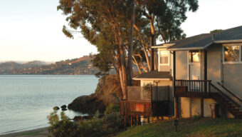 A house with a wooden deck is situated on a grassy slope overlooking a calm body of water in Point San Quentin Village. Tall trees surround the house, and a rocky shoreline is visible in the distance. The sun appears to be setting or rising, casting a warm light over the scene.