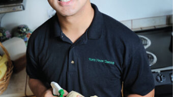 A person stands in a kitchen holding a spray bottle and a yellow cloth. They are wearing glasses and a black polo shirt with text that reads "Nano House Cleaning." The Green Grad poster hangs on the wall, complementing the wooden cabinets, stovetop, and basket with items on the counter.