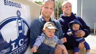 Two bio-dads are each holding a baby, standing beside a tank labeled "LC Biodiesel." The man on the left is wearing sunglasses and both men are smiling. The baby on the left is wearing a denim cap, and the baby on the right is in a blue outfit with a hood.