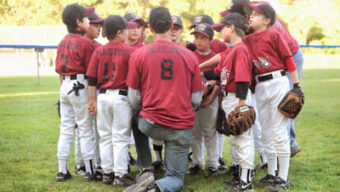 A baseball coach kneels in front of his young team, all wearing maroon jerseys and white pants, as they huddle together on a grassy field. The players, some holding gloves, listen attentively. Trees and a fence are visible in the background... And They Will Come.