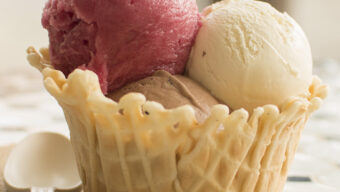 A waffle bowl filled with three delicious scoops of ice cream sits on a napkin atop a tile table. The flavors include raspberry sorbet, vanilla, and chocolate. A white plastic spoon lies to the left of the bowl while the background is softly blurred.