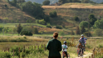 A person runs along a dirt trail in the scenic, hilly landscape of Santa Venetia, wearing athletic clothes. In the distance, two adults and two children ride bicycles on the same path. The area is lush with greenery and rolling hills.