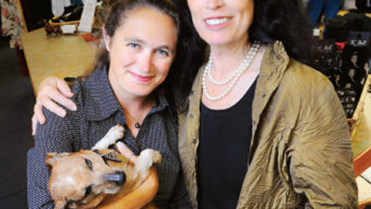 Two women are standing in a clothing store, smiling at the camera. One woman, on the left, is holding a small brown dog. Surrounded by racks of clothes and pictures on the back wall, the atmosphere exudes friendly and casual vibes—quite fitting for some hopeful news in this charming setting.