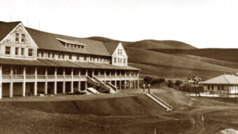 A sepia-toned historical photo of a large building with multiple gabled roofs, a long row of windows, and a wrap-around porch evokes the nostalgia of Radio Days. The building stands on elevated ground with stairs leading up from a dirt path, set against rolling hills in the background.
