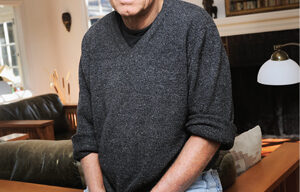 David Harris, a man with light hair and a receding hairline, is standing in a cozy, well-lit room. He wears a dark gray sweater and ripped jeans. Behind him, wooden furniture and a mirror adorn the walls. The space is bathed in both natural light and indoor lighting.
