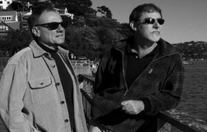 Two men in sunglasses and casual clothing stand on a pier, with one leaning on the railing. They look off into the distance, enjoying the scenic view of a waterfront with houses and trees in the background. This black-and-white image captures the essence of Living South by the water’s edge.