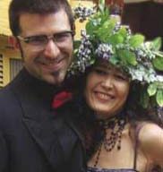 A smiling couple poses together at ArtRageous. The man, with short dark hair, glasses, and a goatee, wears a black suit and red bow tie. The woman has long dark hair and wears a dress adorned with a floral headdress and decorative necklace.