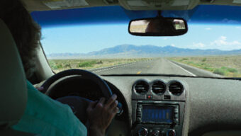 A person is driving a car on a straight, empty road through a vast, flat landscape with mountains visible in the distance. The view is from inside “The Loneliest Hybrid,” looking through the windshield at the driver’s hands on the steering wheel.