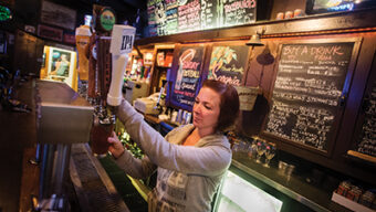 A woman with brown hair wearing a gray hoodie is pulling a draft beer in the dimly lit Smitty's Bar. Behind her are colorful chalkboards displaying various drink options and specials. Shelves with glasses and bottles are visible in the background.
