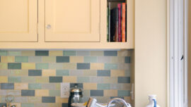 A modern kitchen featuring light yellow cabinets, a small section of open shelving with books, and a colorful tile backsplash in shades of blue and green. The countertop holds a stainless steel sink with a faucet, and a kitchen window is on the right side, showcasing an un-extreme makeover approach.