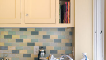 A modern kitchen featuring light yellow cabinets, a small section of open shelving with books, and a colorful tile backsplash in shades of blue and green. The countertop holds a stainless steel sink with a faucet, and a kitchen window is on the right side, showcasing an un-extreme makeover approach.