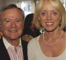 A man and woman, standing closely together at a festival indoors, both smiling at the camera. The man is wearing a dark blazer and light pink shirt, while the woman is in a light-colored top with a pearl necklace. There are other people in the background.