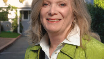 A woman with shoulder-length blonde hair stands outside smiling, wearing a green jacket over a white collared shirt. Seizing the day before her light-colored, two-story house adorned with solar panels and lush greenery, she enjoys the sunny sky and trees in the background. Carpe Diem!