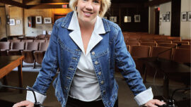 Shawn Marshall, with short blonde hair, is standing and leaning against a dark wooden table in a room with wooden beams and rows of chairs in the background. Wearing a blue denim jacket over a white shirt, they are smiling at the camera.