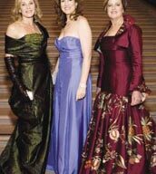 At the Opening Night Gala of the San Francisco Ballet's 75th Anniversary, three women in formal gowns stand side by side on a staircase. The first woman wears a dark green off-the-shoulder dress and long gloves, the second woman is in a strapless light blue gown, and the third woman is in a dark red dress with a floral pattern. They are smiling.