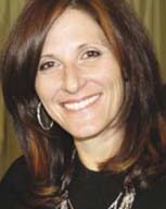 A woman with shoulder-length brown hair and subtle highlights smiles at the camera. She is wearing hoop earrings, a black top with a statement necklace, and stylish shoes. The background is a plain curtain.