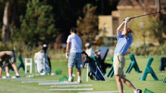 A golfer swings a club on the driving range at Peacock Gap, with several other people practicing in the background. Golf bags and equipment are scattered on the grass, and green trees are visible in the distance. The atmosphere is lively and focused.