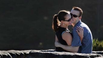 A couple stands outdoors, embracing each other affectionately. The man, wearing sunglasses and a blue checkered shirt, kisses the cheek of the woman, who is also wearing sunglasses and has her hair pulled back. Stone structures are visible in the foreground. Carpe Hug 'Em!