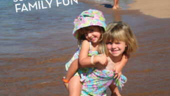 Two young girls playing together on a sunny beach in Hawaii, one giving the other a piggyback ride. The sun shines brightly as people enjoy the water and sandy shore. Palm trees and buildings are visible in the background. Text on the image reads, "SUN-SATIONAL FAMILY FUN.