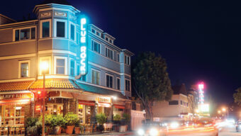 Nighttime street view of a corner building labeled "Blue Rock" illuminated by neon signs. The building, with its decorative architecture, has multiple floors and features a restaurant with outdoor seating on the ground floor. Cars with blurred lights drive past, creating a vibrant Larkspur ambiance.