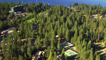 Aerial view of picturesque Incline Village featuring a forested area with scattered buildings and tennis courts, leading to a large, tranquil blue lake. Mountains are visible in the distance under a clear sky.