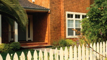 A charming two-story homestead with wooden siding, large windows, and a sloped roof. It is surrounded by green bushes and tall grasses in a picturesque valley, with a white picket fence in the foreground. A leafy tree is on the left side of the house.
