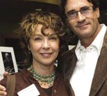 Two people are posing and smiling for the camera at an indoor event. The person on the left, likely attending the Milley Awards, has curly hair and wears a brown top with a lanyard and necklace. The person on the right has short dark hair, glasses, a white shirt, and a brown jacket.