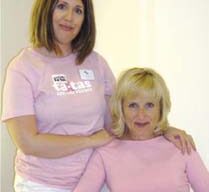 Two women posing together indoors against a neutral background at NH2 Salon. The standing woman, in a light pink t-shirt, has medium-length brown hair, while the seated woman, also in a light pink top, boasts shoulder-length blonde hair. Both look at the camera with relaxed expressions.
