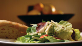 A plate featuring a fresh green salad with lettuce, sliced almonds, and what appears to be bacon, accompanied by a piece of bread. In the background, an AVA bowl of fruit adds a touch of freshness. The scene is warmly lit.