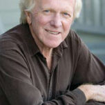 An older man with light-colored hair, wearing a dark long-sleeved shirt, is smiling and leaning on a surface with his arms crossed. The background is neutral and out of focus, likely in an outdoor setting—perhaps contemplating Marin's Next Phase.